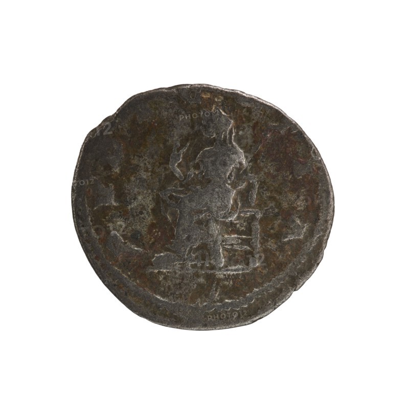 Denarius, Elagabalus reign 218–222 CE. Obverse: IVLIAMAESAAVG — Draped bust of Julia Maesa facing right, pearl border. Reverse: PVDICITIA — Veiled Pudicitia seated on a throne facing left, raising the hem of her veil with her right hand and holding a scepter in her left, pearl border. Roman Empire, struck in Rome, 218–223 CE. Coin; diameter ~17.5–19.0 mm, weight 2.45 g., A round, worn coin with an uneven, slightly irregular edge showing a raised central design of a seated figure facing right. The surface shows mottled areas of dark gray, brown, and hints of dull green and rust, with lighter gray highlights on the raised parts. The figure and surrounding details have a textured, weathered appearance with patches of smoother wear, and the coin sits against a plain white background.