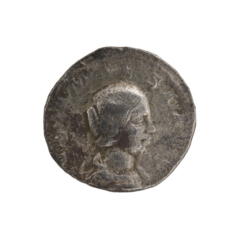 Denarius, Elagabalus 204–222; Roman emperor 218–222. Obverse: IVLIAMAESAAVG — draped bust of Julia Maesa facing right, pearl border. Reverse: PVDIC ITIA — veiled Pudicitia seated on a throne facing left, raising the hem of her veil with her right hand, holding a scepter in her left, pearl border. Date: 218–223, mint: Rome, Roman Empire 27 BC–476. Coin; diameter 17.5–18.0 mm, weight 2.87 g., A worn circular metal object with a raised profile of a person facing right, showing facial features, hair pulled back, and a suggestion of clothing at the neck; the surface is textured and pitted with uneven edges. The visible colors are shades of gray, dark gray, and hints of brownish tarnish.