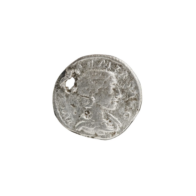 Denarius with hole, Elagabalus reigned 218–222, issuer: Julia Soaemias 180–222; Augusta 218–222. Obv.: IVLIASOAEMIASAVG — Draped bust of Julia Soaemias facing right, pearl border. Rev.: VENVSCAELESTIS — Venus seated on a throne facing left, holding an apple in her right hand and a sceptre in her left; at her feet stands a child with outstretched arms, pearl border. Rome, c. 218–222 CE. Coin; diameter ~17–18 mm; weight 2.35 g., A round, worn metallic object with a rough, pitted surface and an irregular edge, shown against a plain white background; the metal appears silver-gray with darker gray areas in the crevices and lighter silvery highlights on raised portions; the surface shows a faint, worn relief suggesting a profile and ornamental details, plus a small irregular hole near one edge; overall the object looks textured, tarnished, and uneven in color with variations of silver and gray.
