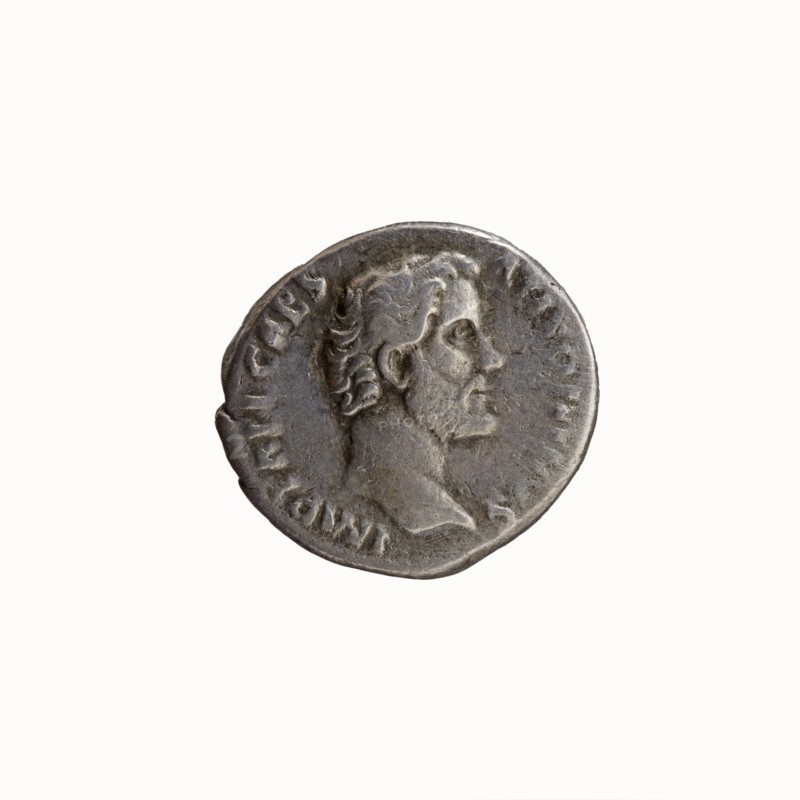 Denarius, Hadrian 76–138; Roman emperor 117–138 and Antoninus Pius 86–161; Roman emperor 138–161. Obverse: Head of Antoninus Pius without a cap to the right, legend IMP T AEL CAES - ANTONINVS around. Reverse: Diana stands to the right, holding a bow in her outstretched left hand and a lowered arrow in her right; legend TRIB PO - T COS around. Iconography: Diana; attributes: bow, arrows. Date: 138. Place of origin: Rome, Roman Empire. Coin type: denarius; diameter ~16.5–18.0 mm; weight 2.88 g; axis 195°., A circular metallic coin with a worn surface showing a raised profile of a bearded man facing to the right, the profile includes detailed hair, ear, eye, nose, lips, and a short neck; the coin has an irregular edge and a textured rim with shallow raised markings around the portrait; visible colors include shades of grey, silver, and darker grey patina across the surface, with subtle lighter highlights on the raised areas.