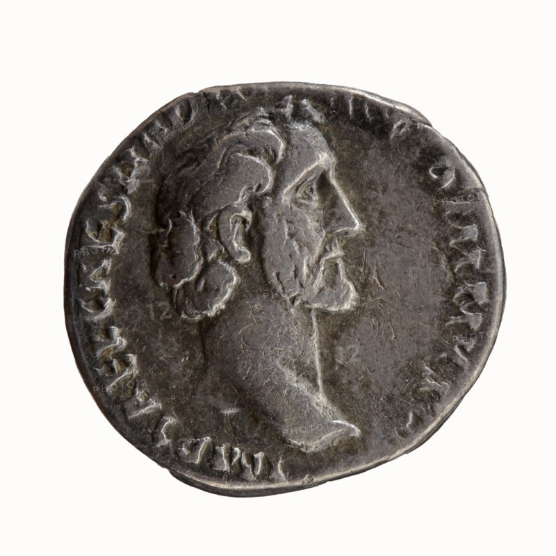 Denarius of Antoninus Pius 86–161; Roman Emperor 138–161. Obverse: Head of Antoninus Pius uncovered to the right, legend IMP T AEL CAES HADRI - ANTONINVS. Reverse: Diana standing to the right, holding a bow in her outstretched left hand and an arrow in her lowered right hand, legend AVG PIVS PM T - RP COS DES II. Iconography: Diana standing; attributes: bow, arrows. Issuer: Antoninus Pius. Date: 138. Place of mint: Rome. Roman Empire 27 BC–476. Coin; denarius. Diameter: 17.0–18.0 mm. Weight: 2.58 g. Axis: 180 degrees., A round metallic coin with a worn surface showing a raised profile of a bearded man facing right; the coin appears mostly dark gray with lighter gray highlights on the raised areas and some subtle brownish tones in recessed parts; a ring of raised decorative marks circles the portrait near the edge; the portrait shows textured hair, an ear, a prominent nose, and a defined neck and chin; the background around the portrait is smoother and slightly darker than the highlight...