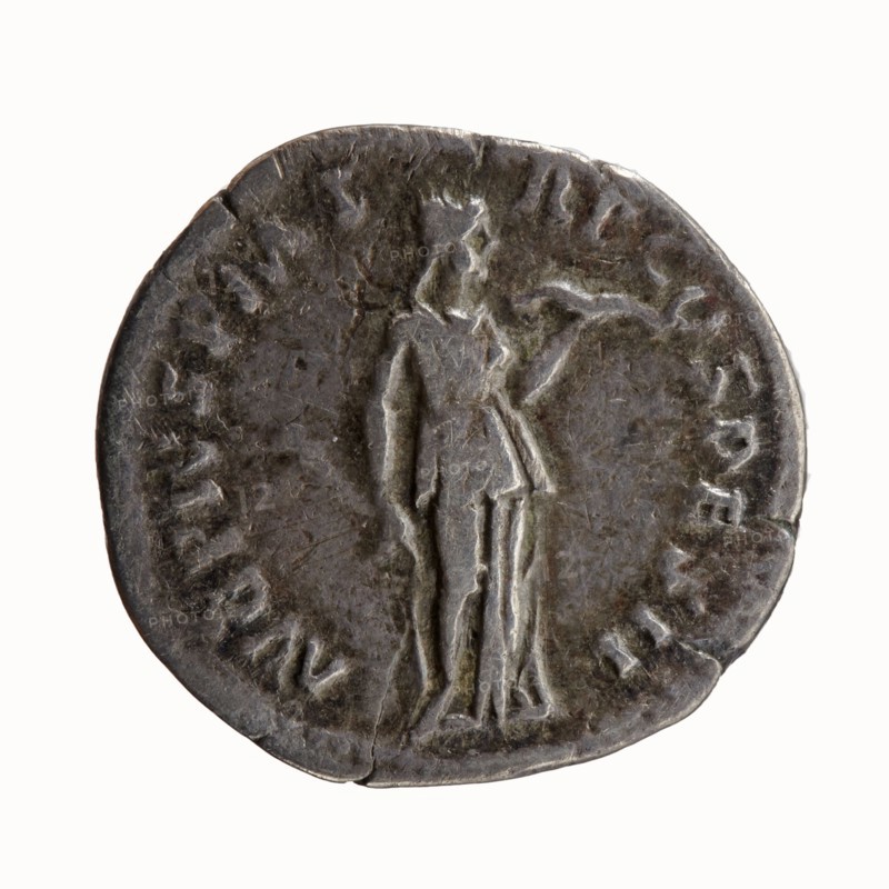 Denarius of Antoninus Pius 86–161; Roman Emperor 138–161. Obverse: head of Antoninus Pius uncovered to the right, legend IMP T AEL CAES HADRI - ANTONINVS. Reverse: Diana standing to the right, holding a bow in her outstretched left hand and an arrow in her lowered right hand, legend AVG PIVS PM T - RP COS DES II. Iconography: Diana standing; attributes: bow, arrows. Date: 138. Place of minting: Rome, Roman Empire. Coin type: silver denarius. Dimensions: diameter ~17–18 mm, weight 2.58 g, axis 180°., A round metallic object with a worn, dark gray surface showing lighter gray highlights, featuring a raised, stylized standing human figure in the center wearing a draped garment and holding an object in one hand; the edges are irregular and slightly dented, and the background shows areas of uneven texture and subtle greenish-brown patina.
