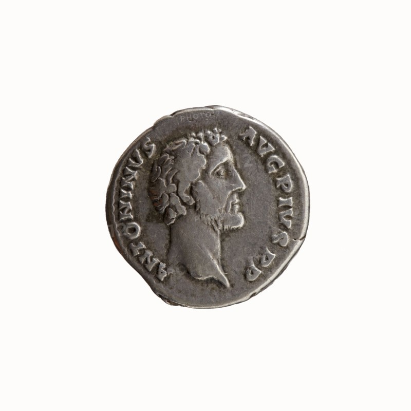 Denarius of Antoninus Pius 86–161; Roman Emperor 138–161. Obverse: Head of Antoninus Pius uncovered, facing right, legend ANTONINVS • AVG • PIVS • PP. Reverse: Container modius with ears of corn and poppy head, legend TR • POT • COS II. Minted Rome, 139. Silver coin; diameter 17.5 mm; weight 3.58 g; axis 165°., A circular metallic coin with a raised profile of a bearded man facing right in the center, surrounded by a raised rim and a ring of small embossed marks; the coin appears silver-gray with darker gray shadows in recessed areas and slightly lighter highlights on raised surfaces, and shows surface wear and minor irregularities along the edge.