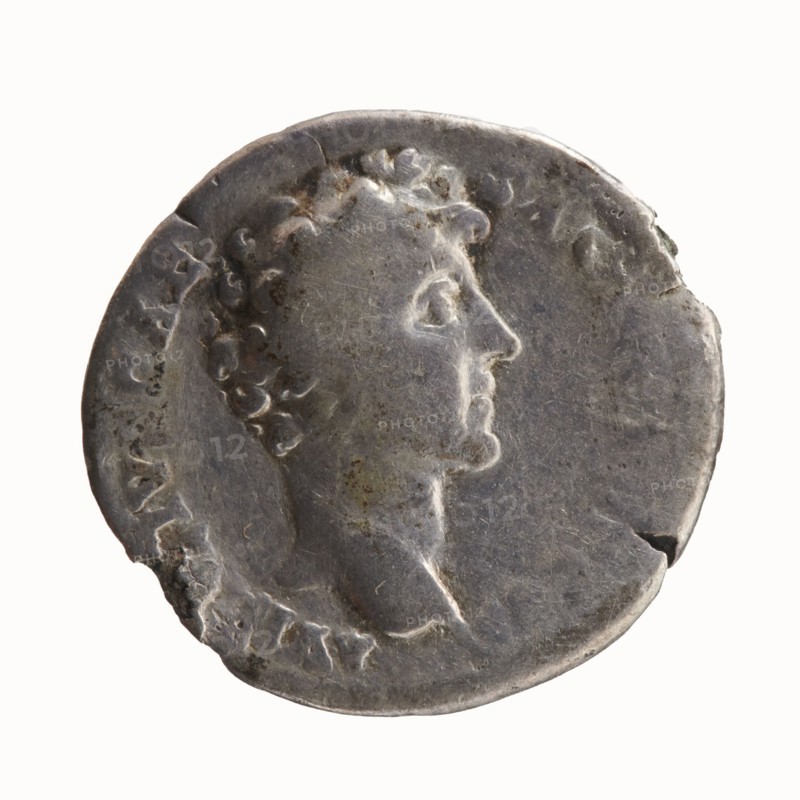 Denarius; obverse: head of Antoninus Pius with laurel wreath facing right, inscription ANTONINVS AVG PI - VS PP TR P COS III around; reverse: head of young Marcus Aurelius without covers right, inscription AVRELIVS CAE - SAR AVG PII F COS around. Issuer: Antoninus Pius 86–161 CE; Roman emperor 138–161 with Marcus Aurelius 121–180 CE; caesar 139–161, heir to the throne. Date: 140 CE. Roman Empire; Rome mint. Coin. Diameters: 16.0 mm, 18.5 mm; weight 2.78 g; axis 225°., A worn circular metallic object with a raised profile of a person's head in right-facing profile, showing curly hair and a visible neck and shoulder; the surface appears aged with scratches, nicks, and small pits, and a few chipped edges; the overall color is silvery gray with areas of darker gray tarnish and small patches of brownish discoloration; the background behind the object is a pale off-white.