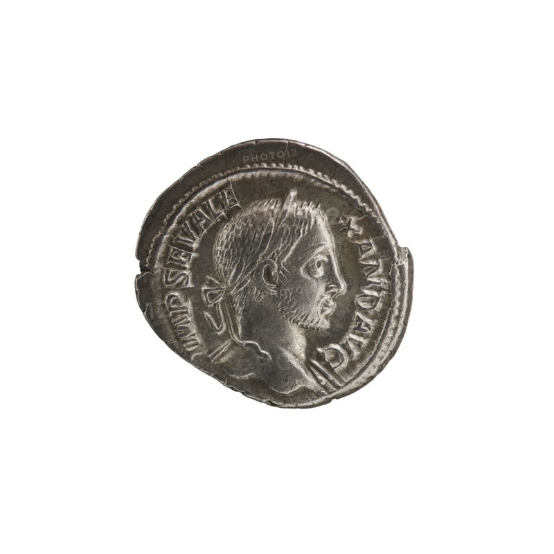 Denarius of Alexander Severus Roman emperor, 222–235. Obverse iconography: standing emperor with spears and globes. Date: 230. Place of origin: Rome, Roman Empire 27 BC–476. Medium: coin. Measurements: diameter 18.0 mm and 20.5 mm; weight 3.99 g., A round metallic coin with a worn surface showing a profile of a man's head facing right, featuring short hair, a tied ribbon at the back, and a short beard; the coin's edge is irregular and slightly damaged in places, and there is raised decorative rim detail around the portrait. The visible colors are silver, gray, and darker gray tones.