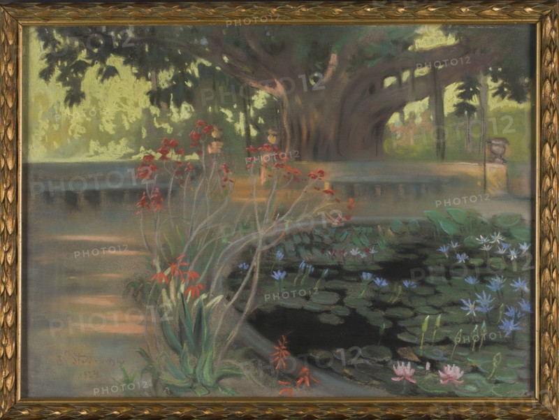 Lotuses in the Park in Palermo, Kazimierz Stabrowski 1869–1929, painter; pastel on paper, 1924; 30.0 × 39.5 cm. Depicts lotus flowers and pond in the botanical gardens Orto botanico of Palermo, Sicily, Italy; water landscape, gardens. Polish artist; Polish pastel., A framed painting showing a garden scene with a large tree in the background, dark brown and muted green trunk and branches, and a canopy of dark green and yellowish leaves. In front of the tree is a low stone wall in gray and beige tones that curves around a pond filled with dark water and many green lily pads. Several water lilies and small flowers in pale blue, light pink, and white float among the pads. Near the pond edge there are clusters of tall, slender plants with green leaves and red and orange blossoms. The ground and surrounding paths show muted shades of gray, brown, and soft peach, while distant foliage includes tones of light yellow-green and olive. The painting is set within an ornate gold frame.