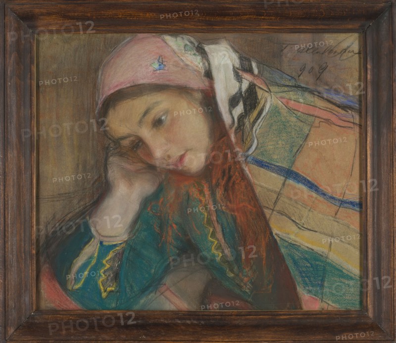 Portrait of a Girl in Krakow Costume, Teodor Axentowicz 1859–1938. Pastel, 1909. Height 33.5 cm; width 44.5 cm. Female portrait of a peasant girl in a Krakow Cracovian costume with scarf headgear; Polish pastel painting in the Young Poland style; ethnographic subject; Krakow., A framed pastel portrait of a young woman leaning her head on her hand, wearing a pink headscarf and a colorful shawl; her face is rendered in soft flesh tones with rosy cheeks and red lips, and her hair shows reddish orange highlights. The shawl and clothing include teal, green, blue, yellow, black, white, peach, beige, and coral tones arranged in striped and patterned sections. The background is a muted brown. The frame is dark wood with visible grain.