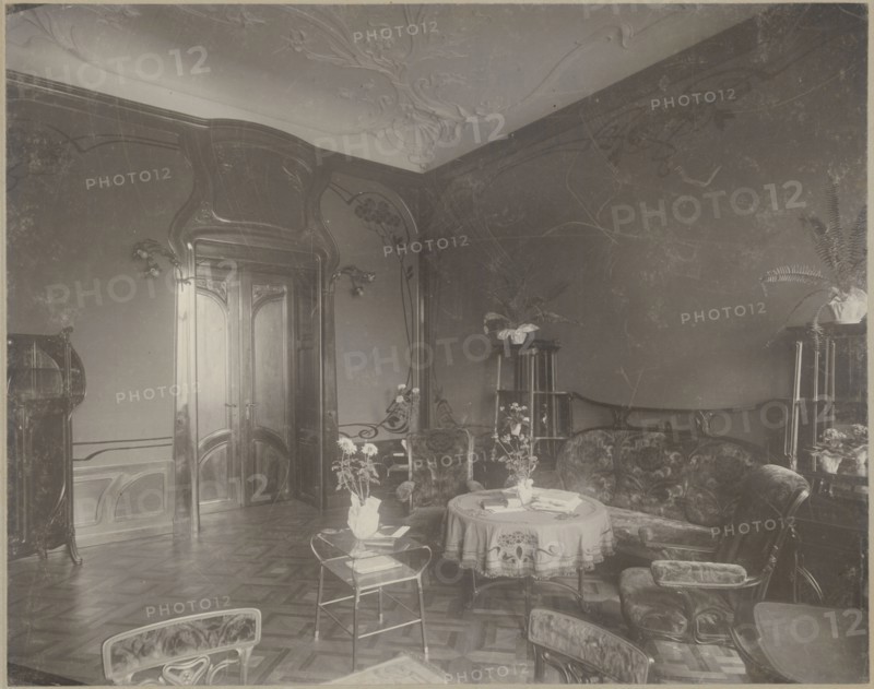 Lódz. Maurycy Poznanski's Palace. View of the drawing room with Art Nouveau decoration and furnishings, Piotrowski photographic studio; 1893–1934 author. Architecture; interiors; palaces; furniture; fabrics; paneling; stucco; doors; Art Nouveau style; modernism; Poland; c. 1900. Photography; photographic print: height 23.5 cm, width 29.6 cm; backing: height 35.1 cm, width 40.6 cm; print glued across the entire surface to decorative cardboard., The image shows an interior room with wooden paneled walls and carved wooden double doors, ornate molded ceiling details, a patterned wooden floor, a curved upholstered sofa and matching armchairs, a round table covered with a decorative cloth and a few items on top, a small metal-framed side table with a potted plant, several potted plants on tall stands, a curved wooden display cabinet, wall-mounted light fixtures, and a few additional chairs visible at the edges; visible colors include shades of brown, beige, cream, and muted green.