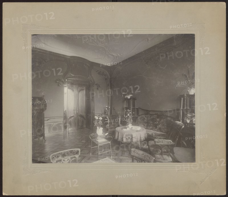 Lódz. Maurycy Poznanski's Palace . View of the drawing room with Art Nouveau decoration and furnishings, Piotrowski photographic studio; 1893–1934 author, architecture, interiors, Polish architecture, palaces, furniture, Lódz Lódz Voivodeship, fabrics, paneling, stucco, doors, Art Nouveau style, Poland culture, modernism style, print glued across the entire surface to decorative cardboard, c. 1900, Lódz place of origin, photography, photographic print: height 23.5 cm, width 29.6 cm; backing: height 35.1 cm, width 40.6 cm, The image shows an interior room with ornate woodwork and carved double doors, a decorative ceiling, patterned wooden floor, and curved walls; there is a group of upholstered chairs and a curved sofa arranged around a round table covered with a cloth, with vases of flowers on the table and on small side tables; several tall plant stands with potted plants are visible near the walls, and a small glass-topped table with a vase sits closer to the foreground; the visible ...