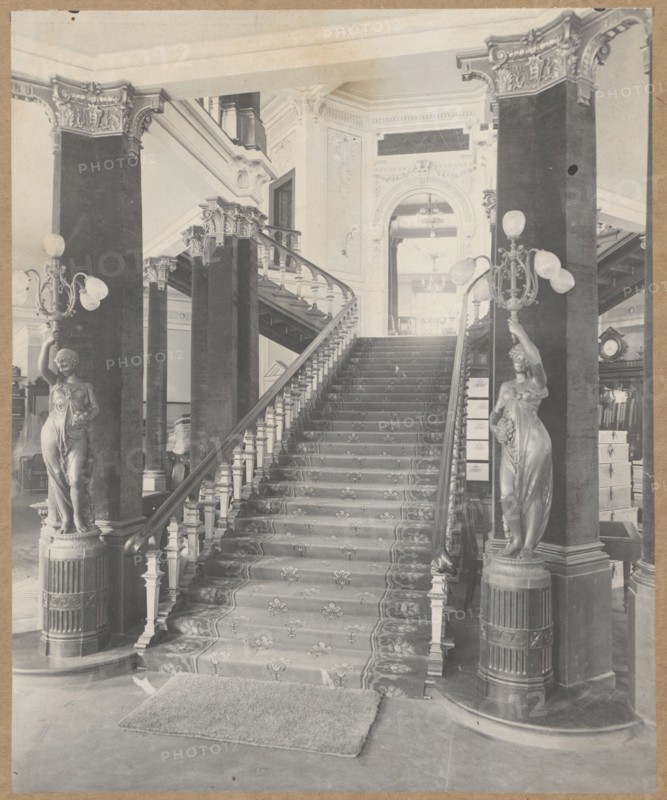 Staircase in the building of the Boguslaw Herse Fashion House at 150 Marszalkowska Street in Warsaw, Boguslaw Herse Fashion House Warsaw; 1868–1936, fashion, interiors, shops, fashion houses, Warsaw Masovian Voivodeship, tenement houses, Art Nouveau, Marszalkowska street, Poland, modernism, print glued across the entire surface onto a museum backing, ca. 1910, photograph from nature, photograph, height 26.7 cm, width 21.8 cm, A grand interior scene showing a wide central staircase with a patterned runner carpet and carved wooden handrails flanked by tall ornate columns. A pair of sculpted female statues stand on decorative pedestals at the base of the staircase, each holding multi-globe light fixtures. The staircase rises to a landing with balustrades and a large arched doorway visible above, with decorative molding and relief work on the walls. There is a rectangular mat at the foot of the stairs on a smooth floor. Visible colors include shades of gray, black, and off-white, with vari...