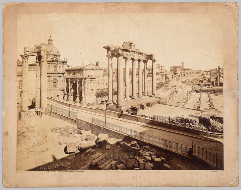 Roma. Foro Romano preso dal Campidoglio, James Anderson 1813–1877, photographer; Anderson Rome, publisher. Subjects: Forum Romanum Rome; architecture—ancient, Italian, foreign; Rome, Italy. Medium: photographic print glued across the entire surface onto thick cardboard backing. Date: ca. 1880. Dimensions: photographic print: 42.1 cm × 55.0 cm; cardboard: 48.9 cm × 62.5 cm., The image shows an open archaeological site with ruined classical columns and stone fragments, with a row of tall standing columns in the center and several shorter columns and ruins scattered around. A road or pathway with metal railings runs through the middle foreground, curving slightly and casting shadows. Buildings with ornate facades and a domed structure appear behind the ruins on the left, and more ruins and distant buildings extend into the background on the right. The ground and stone surfaces are shades of beige and tan, the columns and buildings are light beige, the railings and shadows are darker brown...