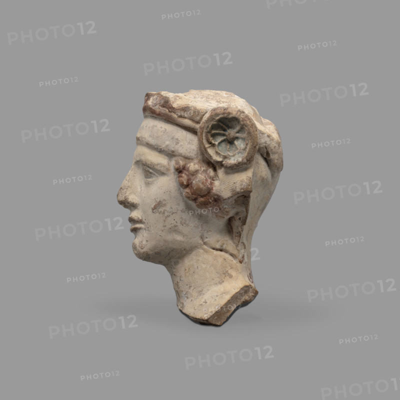 Head of a figurine of Dionysus. Head of a young man with a face devoid of [expression], wide compact mouth, triangularly outlined face with carved cheeks, large eyes, prominent nose and gently outlined eyebrows. The youth wears a diadem decorated with rosettes at the sides and a thick wreath. Traces of red paint (cinnabar) on the hair, lips and diadem; blue in the rosette recesses and on the wreath. 5th–4th century BC, Taranto, Southern Italy, figurine. Height 8.7 cm, width 8.3 cm, depth 6.3 cm, weight 0.28 kg.