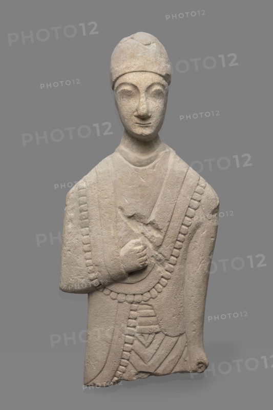 Fragment of a votive figure of a man. The fragment depicts a standing man dressed in a tutulus and himation. The outer edge of the himation is framed by an ornament in the form of semicircular serrated edges. The torso is flattened, the front part is carefully detailed, while the back is flat. The right arm, bent at the elbow, rests on the chest. The right hand is lowered down, close to the torso, the hand clenched into a fist. The face is oval, elongated. The chin is small, the lips are thin, and the nose is long and aquiline. The eyes are slanted and prominent, the eyebrows are arched, and the forehead is low. Conical headdress. Proto-Cypriot Style II, circa 600–480 BC. Cyprus; sculpture. Height 28 cm, width 14 cm.