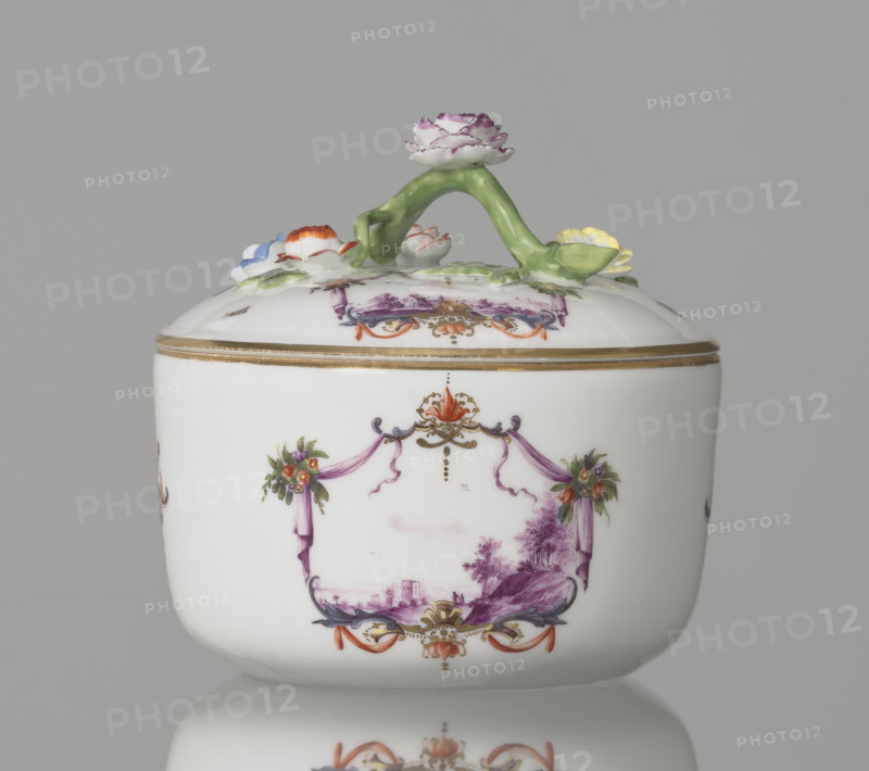 Sugar bowl with a lid and a branch handle, Koenigliche Porzellain Fabrique, Meissen (1710–1763), manufacturer. Cylindrical sugar bowl, rounded at the base, with a slightly arched lid and a plastic, colorfully painted branch handle with four flowers and leaves at the end of the side shoots: three forget-me-nots, two brick-red flowers and one yellow. On the sides of the body two cartouche frames and on the cover one, each framed by two purple draperies suspended from the sides on small flower bouquets and dry rocailles, topped at the top and bottom on the axis of symmetry by brick-red draperies suspended on plant branches. Within the cartouches are landscape miniatures with architecture and staffage painted in purple camaieu: 1) a fragment of a hill with trees, two figures, and a building in a valley; 2) two buildings among bushes; cover: 3) a plateau with a house and a hill in the distance on the right. Between the cartouches on the body are two colorful ornamental-floral motifs; on the...