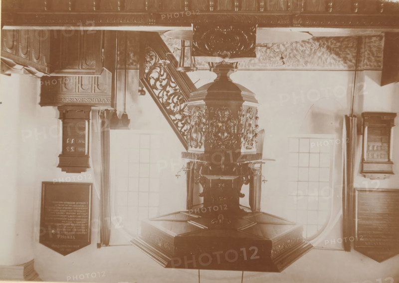Pulpit in the Old St. Victor Church in Batenburg, photograph, page, photographer: Benjamin Charlé (possibly), Batenburg, c. 1903 - in or before 1908, paper, baryta paper, daylight collodion silver print, height 111 mm x width 153 mm, Photo on twelfth unnumbered page., pulpit, ambo, chancel, Oude Sint-Victorkerk, An ornate carved wooden pulpit with decorative panels and a hexagonal base stands beneath a carved wooden staircase with intricate balusters, flanked by tall windows with curtains and framed boards mounted on the walls, all shown in a sepia-toned photograph.