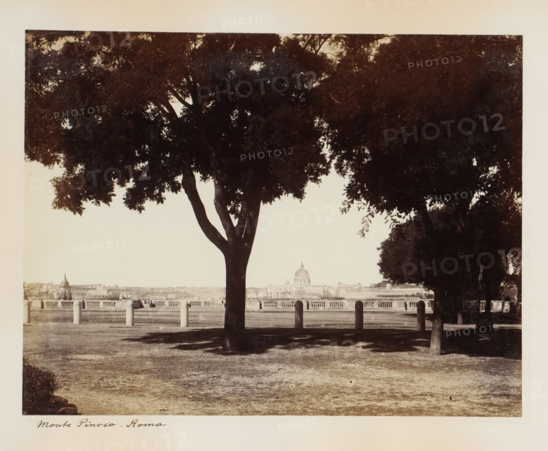 View of Rome from Monte Pincio, Monte Pincio. Rome, album page, photograph, This photo is part of an album., Pincio, 1870 - 1890, photographer, Rome, paper, albumen print, height 202 mm x width 263 mm, garden, city-view in general, 'veduta', trees, public gardens, park, church (exterior), Vatican City, St. Peter's Basilica, A wide, sepia-toned photograph showing a foreground park area with bare, slightly worn ground and shadow patterns, framed by large, leafy trees with dark trunks and dense canopies that create a silhouette against the lighter sky; a row of short stone posts with rounded tops stands between the trees and a balustrade, beyond which an urban riverside panorama is visible with a domed building rising above surrounding structures.