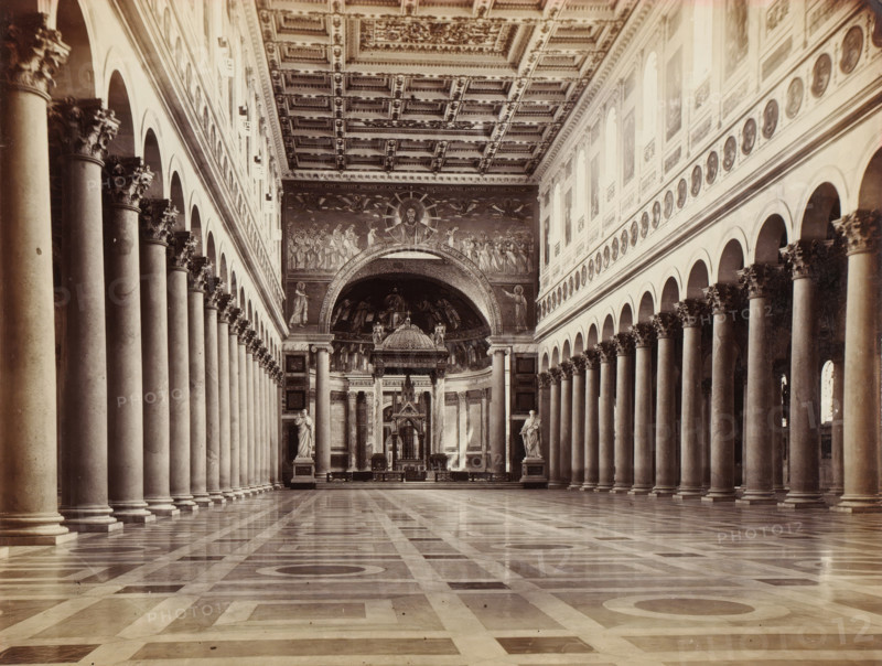 St. Paolo fuori le Mura. Rom, Interior of Saint Paul Outside the Walls in Rome, Italy, album page, photograph, This photo is part of an album., Rome, 1870 - 1890, photographer, paper, albumen print, height 212 mm x width 281 mm, interior of church, Saint Paul Outside the Walls, A wide interior view of a large hall with a long central aisle flanked by rows of tall columns topped with ornate capitals, arched openings above the columns on both sides, a patterned polished floor reflecting light, a richly decorated coffered ceiling, large wall mosaics and paintings near the far end, an arched apse containing sculptural figures and an ornate altar structure, and two freestanding statues positioned near the front of the central space.