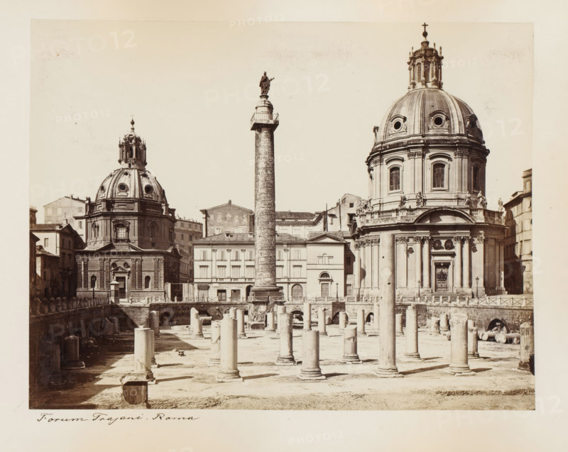View of the Forum of Trajan in Rome, with the Column of Trajan and the churches Santissimo Nome di Maria and Santa Maria di Loreto, Italy, Forum Trajani. Roma, album page, photograph, This photo is part of an album., Rome, 1870 - 1890, photographer, paper, albumen print, height 205 mm x width 265 mm, square, circus, church (exterior), landscape with ruins, column, pillar, architecture, Column of Trajan, Forum of Trajan, Santissimo Nome di Maria al Foro Traiano, Santa Maria di Loreto, A wide sepia photograph of an ancient urban square showing two large domed churches with ornate façades flanking a tall cylindrical column topped by a statue, in front of them a foreground of archaeological ruins with many standing and fallen stone columns and carved blocks, surrounding buildings with multiple windows and tiled roofs visible behind the column, decorative architectural details on the churches including rounded windows and sculpted figures, and the scene framed by a pale border.