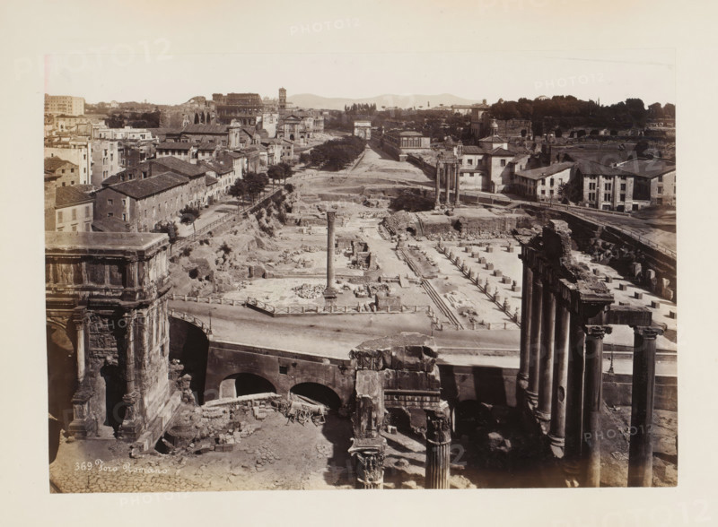 Foro Romano, Forum Romanum in Rome, Italy, album page, photograph, This photo is part of an album., number: '369', Rome, 1870 - 1890, photographer, paper, albumen print, height 188 mm x width 269 mm, market, square, circus, landscape with ruins, Forum Romanum, A wide sepia-toned view of ancient ruins and surrounding buildings showing rows of stone columns and fragmented architectural remains across an excavated area, with several freestanding tall columns visible in the middle ground and a single taller column rising from a rectangular paved area, arched structures and a bridge-like stone construction in the foreground, low multi-story buildings with tiled roofs lining a street on the left, tree-lined paths and sparse vegetation interspersed among the ruins, a range of distant hills along the horizon, and portions of ornate carved stone façades and column capitals visible at the image edges.