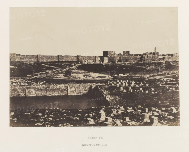 Jerusalem. Birket-Mamillah, Mamilla basin outside the old city wall of Jerusalem, photograph, page, photographer: Auguste Salzmann, Jerusalem, printer (person): Louis-Désiré Blanquart-Evrard, Lille, publisher: Gide et J. Baudry, Paris, 1854 - 1856, printer (person), pasteboard, paper, salted paper print, height 153 mm x width 217 mm, Photo on unnumbered page., city-view in general, 'veduta', pond, pool, Birket-Mamillah, A wide sepia-toned photograph showing a distant stone city wall and clustered buildings along a horizon above a sloping landscape, with a foreground of scattered rectangular stone markers and low ruins, layered terraces and rough ground textures, and a tall narrow tower rising near the right side of the walled structures.