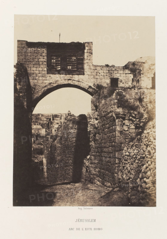 Arch of Ecce Homo, Jerusalem, Jérusalem. Arc de l'Ecce Homo, photograph, page, photographer: Auguste Salzmann, printer (person): Louis-Désiré Blanquart-Evrard, Lille, publisher: Gide et J. Baudry, Paris, 1854 - 1856, Auguste Salzmann, Louis-Désiré Blanquart-Evrard, Gide et J. Baudry, photographer, printer (person), publisher, pasteboard, paper, salted paper print, height 212 mm x width 157 mm, Photo on unnumbered page., gate, entrance, A sepia-toned photograph shows a stone archway set within thick, weathered masonry, with a rectangular upper section featuring a barred window and textured stone blocks; a narrow, shadowed passage runs beneath the arch between rough stone walls that lead toward a distant cluster of low buildings visible through the opening, while rubble and uneven stones line the right side and the ground appears worn and uneven.