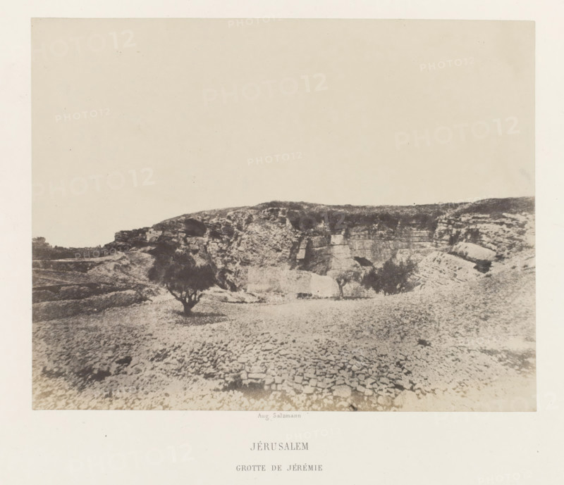 View of the cave of Jeremiah, near Jerusalem, Jérusalem. Grotte de Jérémie, photograph, page, photographer: Auguste Salzmann, Jerusalem, printer (person): Louis-Désiré Blanquart-Evrard, Lille, publisher: Gide et J. Baudry, Paris, 1854 - 1856, printer (person), pasteboard, paper, salted paper print, height 168 mm x width 216 mm, Photo on unnumbered page., cave, grotto, landscape with monument, A sepia-toned photograph showing a rocky landscape with a low cliff or plateau across the midground and a textured foreground of stone and gravel, a few small trees with dark canopies scattered near the base of the cliff, uneven rock formations and ledges along the cliff face, and a large pale sky occupying the upper portion of the image.