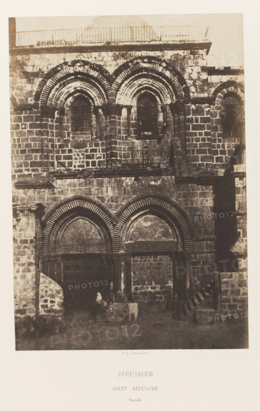 Facade of the Church of the Holy Sepulcher, Jerusalem, Jérusalem. Saint Sepulcre. Façade, photograph, page, photographer: Auguste Salzmann, printer (person): Louis-Désiré Blanquart-Evrard, Lille, publisher: Gide et J. Baudry, Paris, 1854 - 1856, printer (person), pasteboard, paper, salted paper print, height 217 mm x width 151 mm, Photo on unnumbered page., parts of church exterior and annexes: facade, Church of the Holy Sepulcher, A sepia-toned photograph of a stone building façade showing layered arches and rectangular blocks of masonry, with two large rounded archways at ground level and a pair of smaller rounded windows above, decorative concentric ridged molding around the arches, a narrow set of steps visible at one side, rough textured stone surfaces with patches of wear and shadow, and a small indistinct figure near the base of the façade.