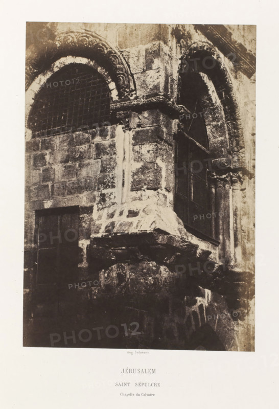 Jerusalem. Saint Sepulcre. Chapelle du Calvaire, Chapel of the Holy Sepulcher, Jerusalem, photograph, page, photographer: Auguste Salzmann, printer (person): Louis-Désiré Blanquart-Evrard, Lille, publisher: Gide et J. Baudry, Paris, 1854 - 1856, printer (person), pasteboard, paper, salted paper print, height 233 mm x width 164 mm, Photo on unnumbered page., architectural detail, parts of church exterior and annexes: chapel, Church of the Holy Sepulchre, A sepia-toned photograph of a stone building façade showing weathered masonry blocks, two arched openings with ornate carved stone surrounds, one arched opening fitted with a metal grid and the other appearing dark and recessed, a projecting stone ledge and a partially visible column with decorative capital, rough textured stones and mortar, and areas of light and shadow across the stone surfaces.