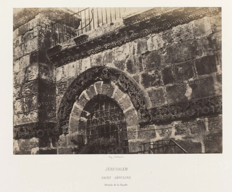Jerusalem. Saint Sepulcre. Détails de la façade, Detail of a window of the Church of the Holy Sepulcher, Jerusalem, photograph, page, photographer: Auguste Salzmann, printer (person): Louis-Désiré Blanquart-Evrard, Lille, publisher: Gide et J. Baudry, Paris, 1854 - 1856, printer (person), pasteboard, paper, salted paper print, height 157 mm x width 224 mm, Photo on unnumbered page., window, parts of church exterior and annexes: facade, Church of the Holy Sepulchre, A close view of a stone facade showing a rounded archway with carved decorative patterns around the arch and a metal grille across the opening, rough textured masonry blocks forming the surrounding wall, a horizontal band of carved ornament above the arch, traces of weathering and discoloration on the stones, and a metal railing visible at the top and lower right edge of the image.