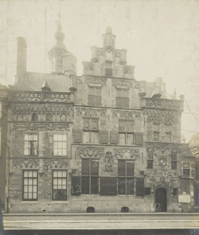 Façade of the Gemeenlandshuis on the Oude Delft in Delft, Gemeentelandshuis. Oude Delft WZ No. 167. Front facade, photograph, inscription: 'Delft. Early 16th century.', inscription: 'The right of reproduction of these photos is reserved by the Publisher.', number: '11', Delft, c. 1900 - c. 1904, photographer, paper, pasteboard, gelatin silver print, gelatin silver developing print, height 156 mm x width 116 mm, Gemeenlandshuis, A sepia-toned photograph of an ornate stone building framed by a dark matte border, showing a multi-story façade with decorative carvings, arched windows with shutters and leaded panes, sculpted reliefs above windows and around a central arched doorway, balustraded rooflines, small dormer structures, and a tower with a bulbous finial rising behind the roof; cobbled street and a canal edge appear in front, and bare tree branches are visible at the sides.