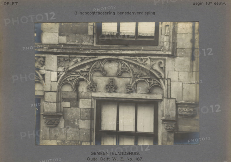 Gemeentelandshuis. Oude Delft WZ No. 167. Blind arch tracery ground floor, Windows of the Gemeenlandshuis on the Oude Delft in Delft decorated with arches and leaf motifs, photograph, number: '14', inscription: 'Delft. Early 16th century.', inscription: 'The right of reproduction of these photos is reserved by the Publisher.', Delft, c. 1900 - c. 1904, photographer, paper, pasteboard, gelatin silver print, gelatin silver developing print, height 117 mm x width 157 mm, ornament derived from plant forms, closed window, Gemeenlandshuis, A grayscale photograph of a stone building facade framed by a darker border, showing a section with ornate carved stonework above and around a window. The carved area features curved arches, floral and foliate motifs, and rounded tracery elements with decorative finials. The surrounding masonry consists of rectangular stone blocks with visible joints and weathering. The window has a dark frame and light-colored curtains or blinds behind the glass. Parts of...