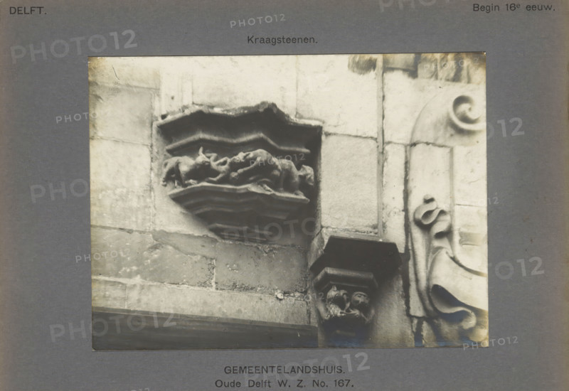 Corrugated stones of the Gemeenlandshuis on the Oude Delft in Delft decorated with fantasy creatures, Gemeentelandshuis. Oude Delft WZ No. 167. Corrugated stones, photograph, number: '16', inscription: 'The right of reproduction of these photos is reserved by the Publisher.', inscription: 'Delft. Early 16th century.', Delft, c. 1900 - c. 1904, photographer, paper, pasteboard, gelatin silver print, height 115 mm x width 155 mm, ornament, hybrid animals, Gemeenlandshuis, A grayscale photograph of a stone facade with carved architectural details showing a recessed decorative shelf with layered zigzag moldings and a central sculpted relief that appears folded and draped, set within large rectangular stone blocks; to the right a vertical column of stone features a rounded carved volute and flowing scroll-like ornamentation, and below the shelf a corbel with two small carved humanlike heads supports another projecting element; the image is bordered by a dark matte with narrow visible edges.