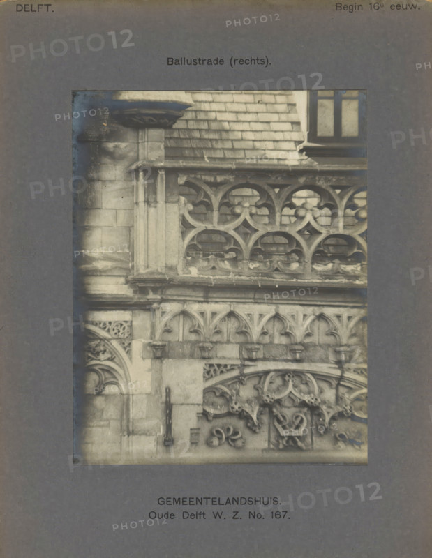 Gemeentelandshuis. Oude Delft WZ No. 167. Balustrade (right), Façade with decorations and a balustrade of the Gemeenlandshuis on the Oude Delft in Delft, photograph, inscription: 'The right of reproduction of these photos is reserved by the Publisher.', number: '19', inscription: 'Delft. Early 16th century.', Delft, c. 1900 - c. 1904, photographer, paper, pasteboard, gelatin silver print, gelatin silver development, height 141 mm x width 111 mm, ornament derived from plant forms, balustrade, Gemeenlandshuis, A close-up photograph of a stone facade showing ornate carved balustrades and decorative arches, with weathered stone blocks and a ledge above the balustrade, a section of tiled roof and a window visible at the top right, and a dark vertical metal hinge or handle affixed to the stone at mid left, all presented within a dark rectangular mat with printed labels above and below the photograph.