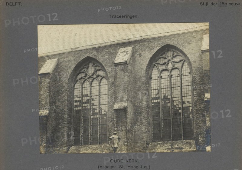 View of the windows of the Old Church in Delft, Old Church. (Formerly St. Hypolitus). Traceries, photograph, inscription: 'The right of reproduction of these photos is reserved by the Publisher.', number: '2', inscription: 'Delft. Style of the 15th century.', Delft, c. 1900 - c. 1904, photographer, paper, pasteboard, gelatin silver print, gelatin silver development, height 116 mm x width 155 mm, parts of church exterior and annexes, Old Church (Delft), A sepia-toned photograph shows a brick church facade with two tall arched stained-glass windows featuring tracery at the tops and rectangular panes below, a steep tiled roof above the wall, small stone projections on the wall near the roofline, weathered bricks with patches of discoloration, and a traditional street lantern mounted on the lower wall between the windows.