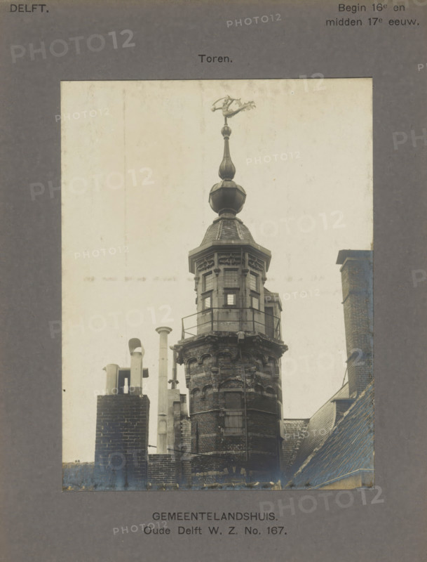 Tower of the Gemeenlandshuis on the Oude Delft in Delft, Gemeentelandshuis. Oude Delft WZ No. 167. Tower, photograph, inscription: 'The right of reproduction of these photos is reserved by the Publisher.', inscription: 'Delft. Early 16th - mid 17th century.', number: '21', Delft, c. 1900 - c. 1904, photographer, paper, pasteboard, gelatin silver print, height 153 mm x width 117 mm, tower (of house or building), weather-vane, Gemeenlandshuis, A sepia-toned photograph of an ornate brick tower rising above rooftops, topped by a decorative cupola with a rounded finial and a weather vane shaped like an animal; the tower has a small octagonal upper section with multi-pane windows and a surrounding metal railing, beneath which the brickwork shows decorative masonry and a narrow vertical window; in the foreground there are several chimney stacks and metal pipes of varying heights on adjacent roof structures, and sloped tiled roofs frame the lower right and left edges of the image.