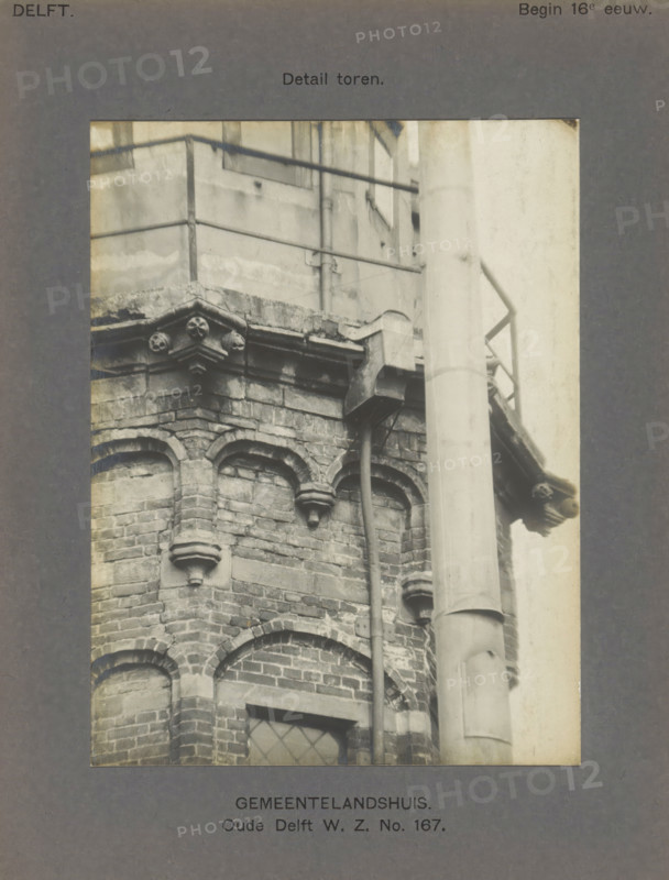 Detail of the tower of the Gemeenlandshuis on the Oude Delft in Delft, Gemeentelandshuis. Oude Delft WZ No. 167. Detail tower, photograph, number: '22', inscription: 'Delft. Early 16th century.', inscription: 'The right of reproduction of these photos is reserved by the Publisher.', Delft, c. 1900 - c. 1904, photographer, paper, pasteboard, gelatin silver print, height 155 mm x width 117 mm, tower (of house or building), Gemeenlandshuis, A close view of a brick corner of a building showing decorative arched brickwork and carved stone corbels beneath a projecting stone ledge, with a vertical drainpipe running down beside the masonry and a large cylindrical column in front, and a metal railing and wall visible above the ledge.