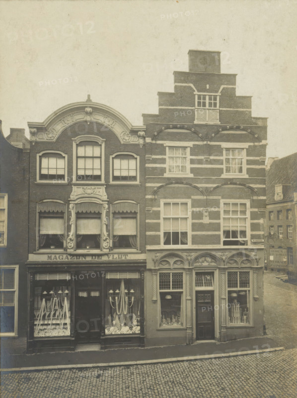 View of two houses and shops on the Wijnhaven in Delft, Wijnhaven No. 15 and No. 16, photograph, inscription: 'Delft. A No. 15. Louis XIV. B No. 16. Early 16th century', number: '24', inscription: 'The right of reproduction of these photos is reserved by the Publisher.', Delft, c. 1900 - c. 1904, photographer, paper, pasteboard, gelatin silver print, gelatin silver developing print, height 155 mm x width 115 mm, shop-window, show-window, A faded photograph shows two adjacent narrow brick buildings with ornate facades facing a cobbled street, the left building featuring a curved decorative gable and three tall windows on the upper floor above a shopfront with display windows and a central door, the right building displaying a stepped gable with horizontal light bands, multiple rectangular windows with small panes, and an arched decorative row above the upper shop windows, while a glimpse of another building and a sloped street appears to the right and a plain frame surrounds the image.