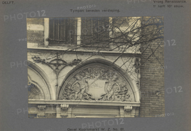 Facade Koornmarkt WZ No. 81. Tympan on the ground floor, Tympanum on the facade of a house on the Koornmarkt in Delft, decorated with two people next to a coat of arms, photograph, inscription: 'The right of reproduction of these photos is reserved by the Publisher.', inscription: 'Delft. Early Renaissance. 1st half of the 16th century.', number: '28', Delft, c. 1900 - c. 1904, photographer, paper, pasteboard, gelatin silver print, developed gelatin silver print, height 115 mm x width 157 mm, urban housing, coat of arms (as symbol of the state, etc.), ornament derived from architectural details, Koornmarkt, A close view of a carved stone facade showing an arched decorative panel with sculpted cherubic figures and ornate floral scrollwork around a central blank shield, framed by curved moldings and supported by carved pilasters with acanthus capitals, with bare tree branches crossing over the stonework and a brick wall visible to the right.