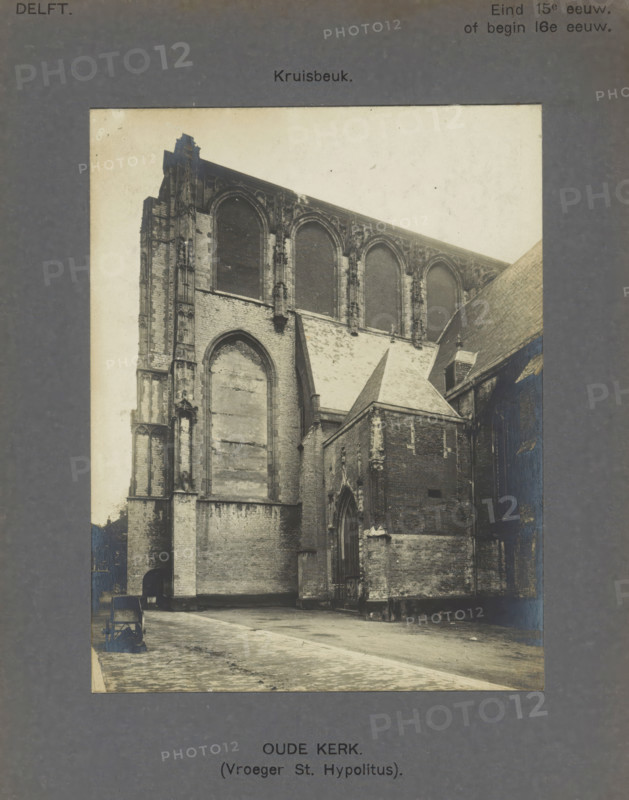 Old Church. (Formerly St. Hypolitus). Transept, View of the transept of the Old Church in Delft, photograph, inscription: 'The right of reproduction of these photos is reserved by the Publisher.', inscription: 'Delft. Late 15th century. or early 16th century.', number: '3', Delft, c. 1900 - c. 1904, photographer, paper, pasteboard, gelatin silver print, gelatin silver developing print, height 150 mm x width 117 mm, parts of church exterior and annexes: transept, Old Church (Delft), A sepia-toned photograph shows a large stone church exterior with tall, arched window openings framed by ornate vertical stonework and a steep sloping roof section; an adjoining lower brick structure with a pitched roof and an arched doorway is attached to the taller wall, and decorative buttresses and carved stone details are visible along the walls; the foreground shows a paved open area with a small cart or wheeled object near the left edge and a narrow path leading toward the building, while the image is...