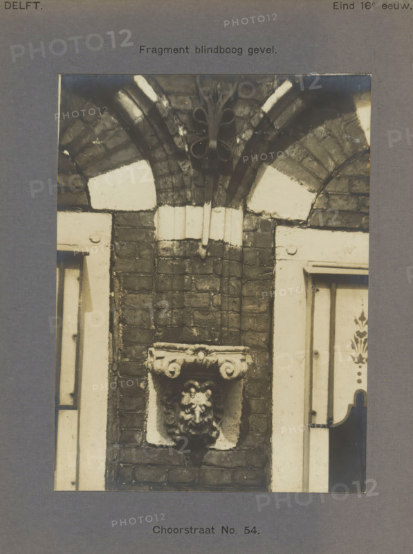 Corb stone decorated with a head in the facade of a building in the Choorstraat in Delft, Choorstraat No. 54. Facade, photograph, inscription: 'The right of reproduction of these photos is reserved by the Publisher.', inscription: 'Delft. Late 16th century.', number: '36', Delft, c. 1900 - c. 1904, photographer, paper, pasteboard, gelatin silver print, developed gelatin silver print, height 157 mm x width 117 mm, urban housing, ornament derived from architectural details, A cropped photograph of a brick wall section showing a decorative stone corbel with a carved face mounted in a recessed square panel, flanked by lighter painted door or window frames on each side; above the corbel the brickwork rises beneath a semi-circular arch with alternating dark and light stone segments and a small carved foliate boss at the arch apex.