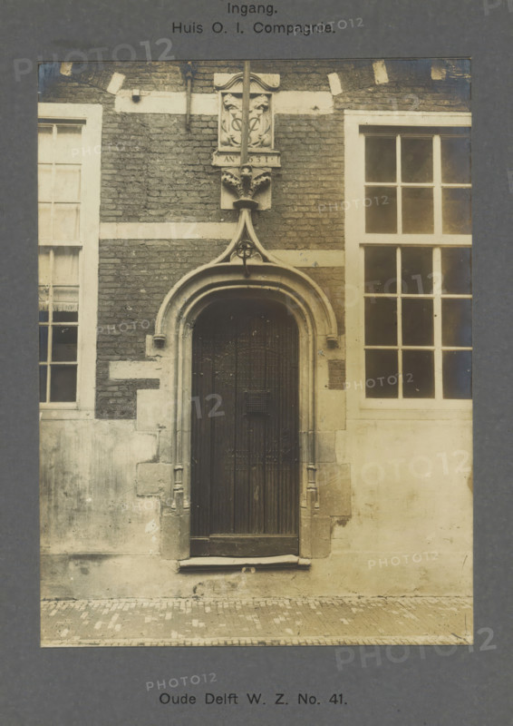 Oude Delft WZ No. 41. Entrance. House of the Dutch East India Company, Entrance to the East India House on Oude Delft in Delft, photograph, inscription: 'The right of reproduction of these photos is reserved by the Publisher.', number: '39', inscription: 'Delft. Early 16th century. Gable stone 1631', Delft, c. 1900 - c. 1904, photographer, paper, pasteboard, gelatin silver print, height 157 mm x width 117 mm, gate, entrance, Oude Delft, A tall arched wooden door set into a stone and brick facade flanked by two tall multi-pane windows, the doorway framed by molded stone with decorative carved elements above including a small ornate plaque and a vertical stone finial, weathered bricks and lighter stone bands surrounding the opening, worn stone steps and a cobbled street in front, and subtle mottling and stains on the lower wall surfaces.