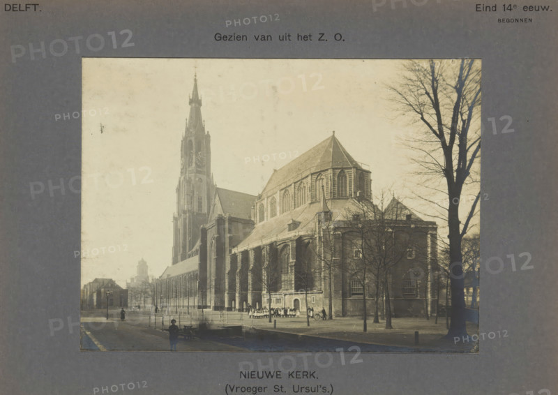 Nieuwe Kerk. (Formerly St. Ursul's). Viewed from the SE, View of the Nieuwe Kerk in Delft with a group of children in front of it, photograph, inscription: 'Delft. Begun late 14th century', number: '4', inscription: 'The right of reproduction of these photos is reserved by the Publisher.', Delft, c. 1900 - c. 1904, photographer, paper, pasteboard, gelatin silver print, height 113 mm x width 154 mm, church (exterior), parts of church exterior and annexes: tower, Nieuwe Kerk (Delft), A sepia-toned photograph shows a large Gothic church with a tall ornate tower and a steeply pitched roof, flanked by leafless trees in the foreground; people stand near the church entrance and along the sidewalk, with a cyclist visible nearby, while a canal or waterway runs along the street where a few pedestrians and a person near a small boat are present.