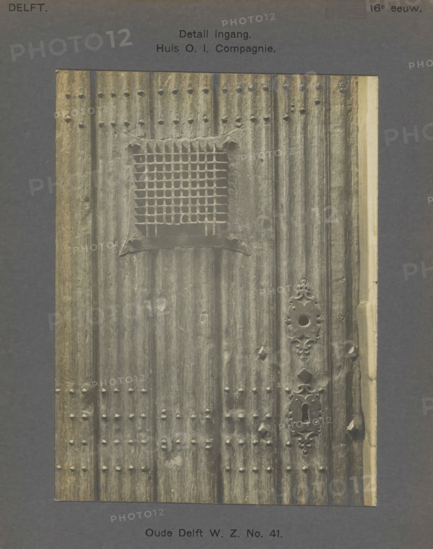 Detail of the entrance to the East India House on Oude Delft in Delft, Oude Delft WZ No. 41. Entrance detail. House OI Company, photograph, inscription: 'The right of reproduction of these photos is reserved by the Publisher.', inscription: 'Delft. 16th century', number: '40', Delft, c. 1900 - c. 1904, photographer, paper, pasteboard, gelatin silver print, height 160 mm x width 120 mm, gate, entrance, lock, door, Oude Delft, A close view of a vertically ribbed wooden door with a square grid-covered opening near the upper center and rows of small rounded studs arranged in horizontal bands, the door surface showing weathered texture and vertical grain. An ornate metal escutcheon plate with a circular hole and decorative flourishes is visible along the right side, and a keyhole plate with an elongated key slot is positioned below it. The door edges show lighter trim on the right and darker worn wood on the left, and the whole image is set against a dark mat background.