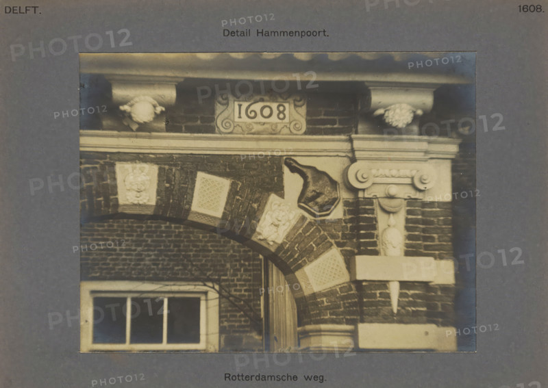 Rotterdamseweg. Detail Hammenpoort, Decoration of a ham on the Hammenpoort on the Rotterdamseweg in Delft, photograph, inscription: 'Delft. 1608', number: '42', inscription: 'The right of reproduction of these photos is reserved by the Publisher.', Delft, c. 1900 - c. 1904, photographer, paper, pasteboard, gelatin silver print, height 118 mm x width 155 mm, gate, entrance, ham, A sepia-toned photograph shows a portion of a brick archway with alternating brick and lighter stone blocks, each lighter block featuring a raised decorative pattern. Above the arch is a stone entablature with molded cornice and ornamental corbels, and a carved plaque set into the brickwork between the corbels. To the right of the arch a stone pilaster with scroll-like molding and bead detailing supports the entablature. A protruding metal bell-shaped object is mounted on the wall above one of the lighter blocks. Below the arch a window with divided panes is partially visible through the opening, and bare branch...