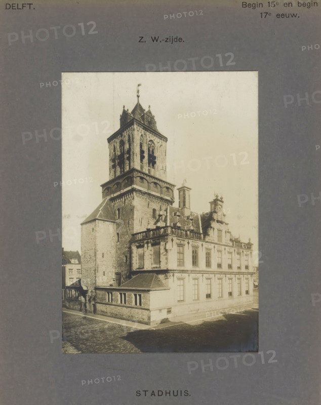 View of the town hall in Delft, Town Hall. SW side, photograph, inscription: 'Delft. Early 15th and early 17th century.', inscription: 'The right of reproduction of these photos is reserved by the Publisher.', number: '44', Delft, c. 1900 - c. 1904, photographer, paper, pasteboard, gelatin silver print, height 138 mm x width 96 mm, town hall, Stadhuis (Delft), A sepia photograph shows a large historic stone building with a tall square tower rising above ornate rooftop structures; the tower has arched openings and a steep roof with decorative finials and a small flagpole, while the adjoining building features rows of rectangular windows, sculpted cornices, and a balustraded roofline with small dormers and chimneys; a cobblestone street runs alongside the building and a low stone wall and entranceway are visible at the corner, with a shadow cast across part of the street and faint outlines of additional buildings in the background.