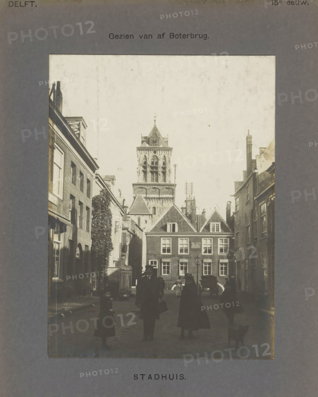 View of the town hall in Delft, seen from the Boterbrug with people, a coach and a dog, Town Hall. Seen from Boterbrug, photograph, inscription: 'Delft. 15th century.', inscription: 'The right of reproduction of these photos is reserved by the Publisher.', number: '45', Delft, c. 1900 - c. 1904, photographer, paper, pasteboard, gelatin silver print, height 155 mm x width 115 mm, dog, bus, coach, townhall, Town Hall (Delft), A narrow street scene framed by a dark border shows tall brick buildings on both sides leading to a square with a gabled building and a large clock tower rising behind it; silhouettes of people in dark clothing stand and walk in the foreground, one adult figure carrying a bag, a child nearby, and a small dog at the edge; windows, chimneys, rooflines, and a lamppost are visible, along with a horse-drawn carriage near the square and architectural details on the tower.