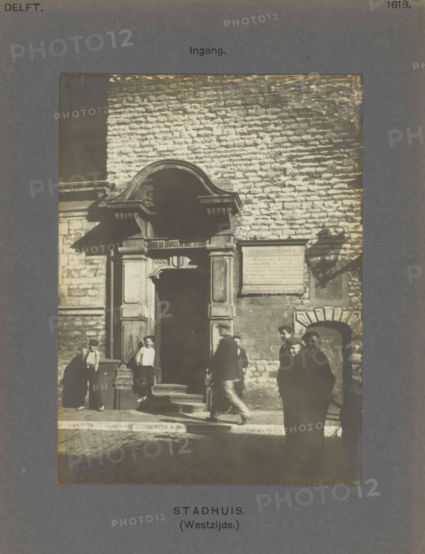 Entrance to the town hall in Delft with boys in front of the gate, Town Hall (West side.) Entrance, photograph, inscription: 'The right of reproduction of these photos is reserved by the Publisher.', number: '46', inscription: 'Delft. 1618.', Delft, c. 1900 - c. 1904, photographer, paper, pasteboard, gelatin silver print, height 155 mm x width 115 mm, town hall, gate, entrance, A sepia photograph shows a stone building facade with a large arched doorway framed by decorative masonry and a smaller arched opening to the side, people standing and walking on the steps and street in front of the entrance, a rectangular sign mounted on the wall beside the doorway, rough textured brickwork covering the wall above, strong sunlight casting distinct shadows, and a dark border surrounding the image.