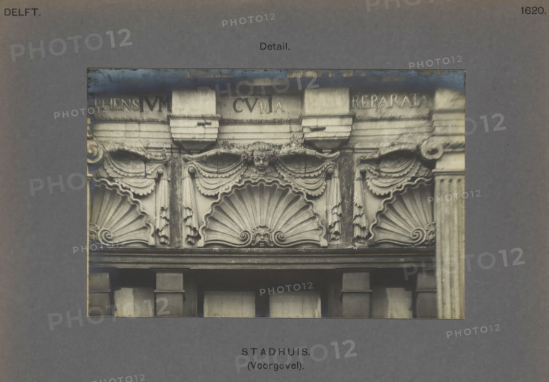 Town Hall (Facade). Detail, Detail of the façade of the town hall in Delft, decorated with shells and cherubs, photograph, inscription: 'Delft. 1620.', number: '48', inscription: 'The right of reproduction of these photos is reserved by the Publisher.', Delft, c. 1900 - c. 1904, photographer, paper, pasteboard, gelatin silver print, height 97 mm x width 148 mm, townhall, cherubs, i.e. children's heads with wings, scallop, shell, ornament - AA - stylized, Town Hall (Delft), A rectangular photograph shows a detailed stone facade with decorative sculpted elements including three large scallop shell motifs, draped garlands, and carved pilasters; a small carved face is centered above the middle shell, flanked by ornate scrolls and hanging tassel-like decorations; the stone appears weathered with darker staining in places and a horizontal cornice runs above the decorative band; the photo is mounted on a gray board with a wide border surrounding it.
