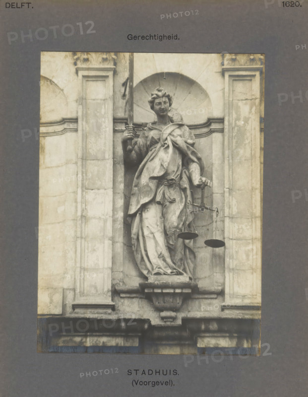 Sculpture of Lady Justice on the town hall in Delft, Town Hall (Facade). Justice, photograph, inscription: 'The right of reproduction of these photos is reserved by the Publisher.', inscription: 'Delft. 1620.', number: '49', Delft, c. 1900 - c. 1904, photographer, paper, pasteboard, gelatin silver print, height 156 mm x width 116 mm, town hall, 'Giudice' (Ripa), A grayscale photograph of a sculpted female figure set into an architectural niche of a stone facade, the figure draped in flowing garments with one hand raised holding a vertical object and the other hand extended slightly downward holding a set of suspended pans, the niche framed by rectangular pilasters and decorative molding, the statue standing on a carved pedestal above a projecting cornice, with parts of the weathered stone surface showing discoloration and surface texture.