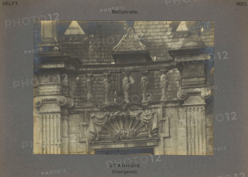 Balustrade on the town hall in Delft, Town Hall (Facade). Balustrade, photograph, inscription: 'The right of reproduction of these photos is reserved by the Publisher.', number: '50', inscription: 'Delft. 1620.', Delft, c. 1900 - c. 1904, photographer, paper, pasteboard, gelatin silver print, height 115 mm x width 156 mm, town hall, balustrade, A photographic detail of a stone façade showing architectural ornamentation including balusters, carved pilasters with fluted shafts and decorated capitals, a broken pediment with draped swags, a scallop shell motif with a central carved face, ornate scrolls and molded cornices, a tiled roof area above with decorative stone elements and a small window opening partially obscured by the balustrade, all presented within a wider neutral-toned mounting.